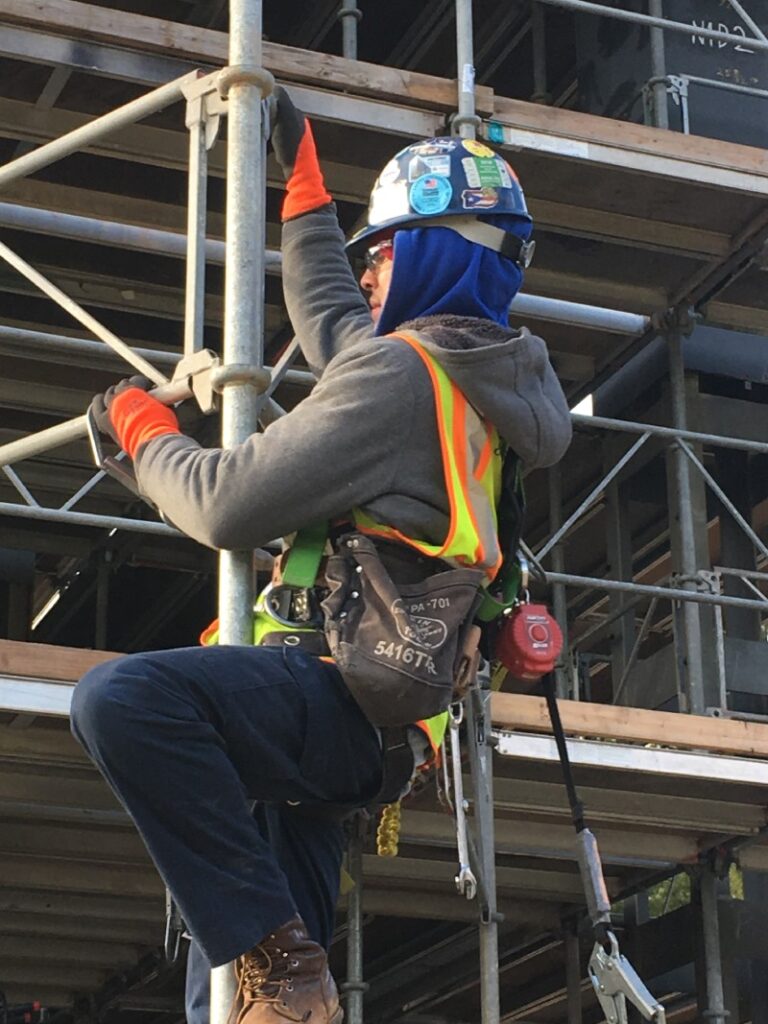 Worker wearing safety gear and harness climbing scaffolding during a building maintenance project.