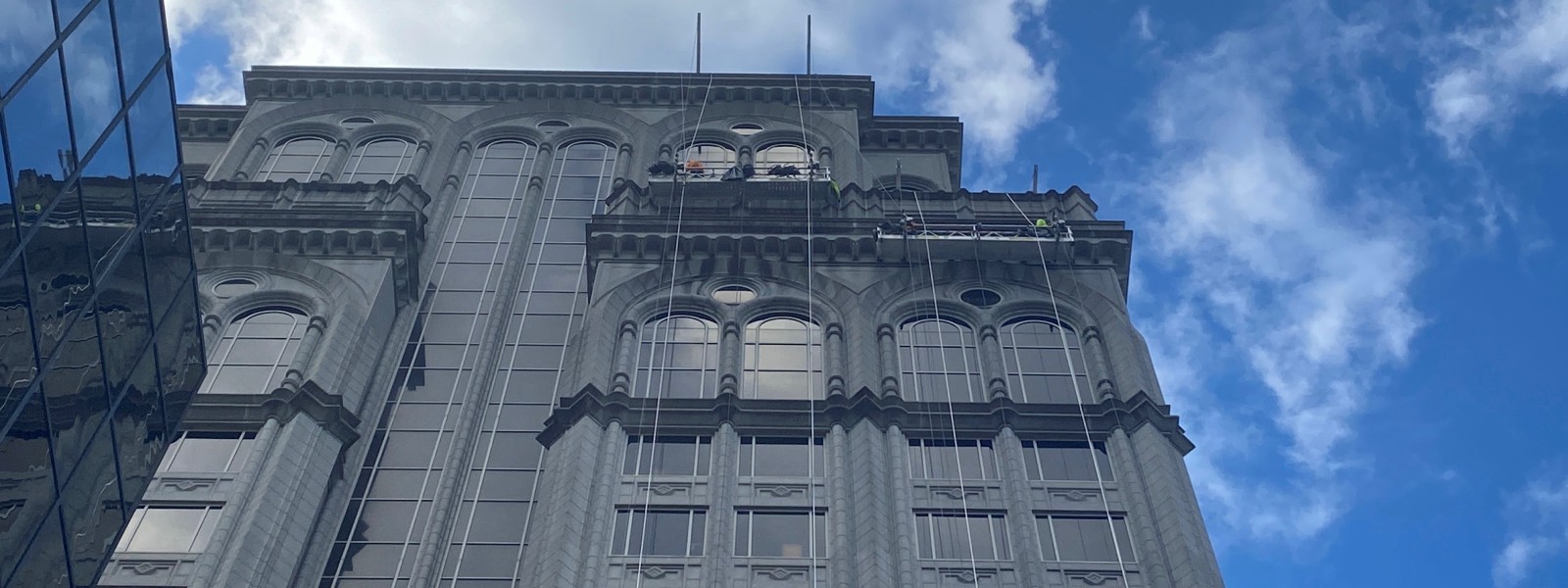 Workers using a suspended swing stage for exterior maintenance on a high-rise commercial building against a bright blue sky.